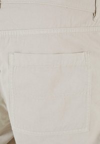 Gaiši bēša audums ar kabatu aizmugurē, kurā ir horizontālas šuves. Materiāls šķiet izturīgs ar gludu tekstūru.