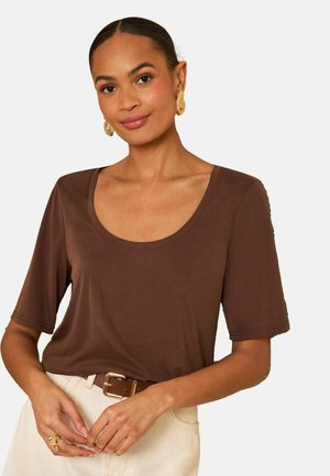 Naine, kes kannab pruuni madala kaelusega lühikeste varrukatega pluusi, beeže pükse pruunist vööst ja kuldseid rõngaskõrvarõngaid, käed õrnalt ees risti.