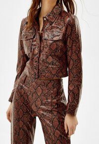 Veste cintrée, brillante, à motif serpent dans des nuances de rouge et noir, avec deux poches poitrine et une fermeture à boutons ; associée à un pantalon assorti.