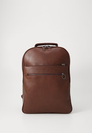 Mochila de cuero marrón con forma redondeada, que presenta un bolsillo delantero con cremallera, asas dobles en la parte superior y una textura suave. Acentos de hardware en plata.