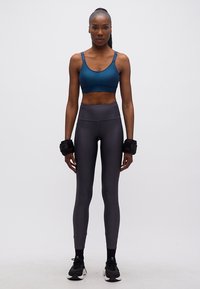 Γυναίκα που στέκεται με πρόσωπο προς τα εμπρός, φοράει μπλε αθλητικό σουτιέν, σκούρο κολάν, μαύρα αθλητικά παπούτσια και βάρη καρπού, σε λευκό φόντο.