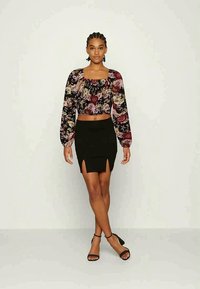 Top cropped preto com padrão floral, mangas longas balão, combinado com uma saia preta de cintura alta com fendas laterais, e sandálias pretas de tiras.
