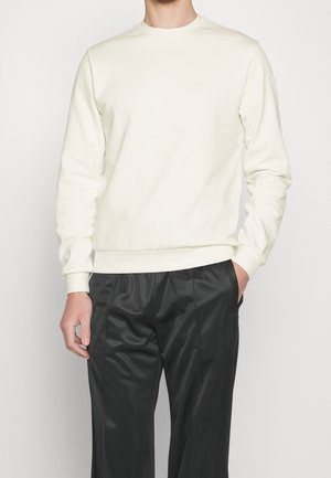 Mand iført en simpel hvid langærmet sweatshirt med ribstrikkede manchetter og linning, kombineret med løstsiddende sorte bukser.