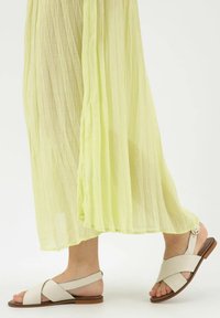 Lichtgele, geplooide rok valt over de voet. Beige leren sandalen hebben een kruisbandontwerp en een platte zool, met een cirkelvormige gesp als accent.