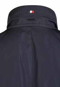 Col montant de veste de pluie marine avec le logo noir "HECHTER" et un petit accent rouge, blanc et bleu. Tissu lisse et texturé.
