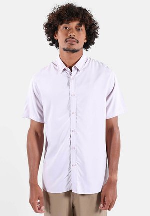 Homme aux cheveux bouclés portant une chemise rose clair à manches courtes avec des boutons et un pantalon beige, debout devant un fond blanc uni.