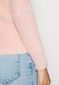 Maglione rosa peloso con maniche lunghe, caratterizzato da un orlo a coste. Abbinato a jeans azzurri chiari, che mette in risalto i dettagli della texture e della forma.