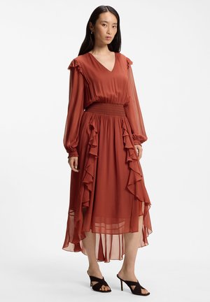 Mujer con cabello largo y oscuro que lleva un vestido de manga larga color óxido con volantes, cintura fruncida, dobladillo asimétrico y sandalias negras de tacón.