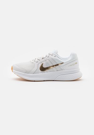 Weißer Nike Laufschuh mit Mesh-Obermaterial, goldfarbenem metallischem Swoosh, gedämpfter Sohle und Gummi-Außensohle, seitlich auf einfarbigem Hintergrund gezeigt.