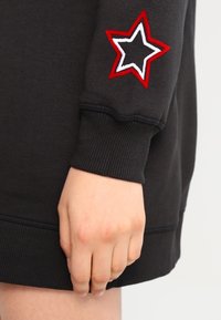 Musta collegepaita ribbivarsilla, jossa on punainen ja valkoinen kirjailtu tähti vasemmassa hihassa. Sujuva kankaan rakenne.