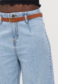 Um par de jeans de denim azul claro apresenta uma cintura alta, corte de perna reta e um cinto de couro bege com uma fivela redonda dourada.