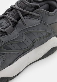 Grå sneaker med en design i blandade material, inklusive mocka och gummidetaljer. Framträdande detaljer är texturerade paneler och en vadderad sula.