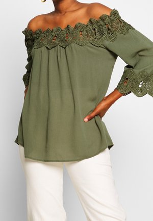 Blusa verde oliva a spalle scoperte con dettagli in pizzo lungo il colletto e le maniche, realizzata in un tessuto leggero, abbinata a pantaloni bianchi.