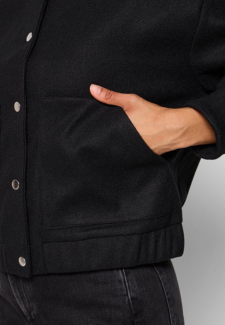 Schwarze Jacke aus strukturiertem Stoff mit silbernen Druckknöpfen und seitlichen Taschen, die eine strukturierte Silhouette und einen gerippten Saum aufweist.