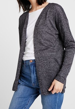 Femme portant un cardigan gris foncé ouvert devant, un t-shirt blanc et un jean bleu, une main reposant près de la hanche.
