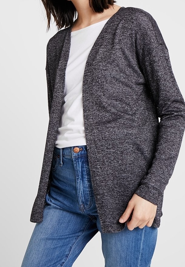 Femme portant un cardigan gris foncé ouvert devant, un t-shirt blanc et un jean bleu, une main reposant près de la hanche.