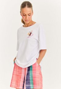 Žena v bílém tričku s grafikou a textem na hrudi a barevnými pruhovanými šortkami, s rukama v kapsách, dívající se dolů.