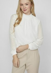 Femme blonde portant un chemisier blanc à manches longues texturé avec des poignets et un col à volants, associé à un pantalon taille haute beige.