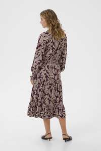 Langes lila Kleid mit einem cremefarbenen Paisleymuster. Verfügt über lange Ärmel, einen elastischen Bund und einen gestuften Rüschensaum. Getragen mit schwarzen Absatzsandalen.