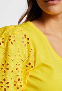 Gulblus med puffärmar som har intrikata blommiga hålmönster, en slät kropp och design med V-ringning.