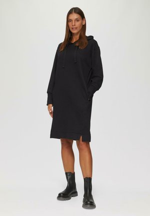 Zwarte hoodie jurk met lange mouwen, zijzakken en een lichtgebogen zoom. Gedragen met zwarte enkellaarzen. Gladde textuur.