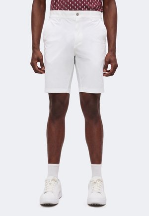 Homme portant un short blanc aux genoux, des chaussettes blanches et des baskets blanches, les mains détendues le long du corps.