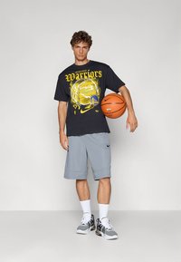 Svart grafisk t-skjorte med gul "Warriors"-trykk, grå shorts og hvite sokker. Holder en oransje basketball, iført grå og svarte joggesko.