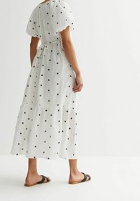 Weißes Maxikleid mit schwarzer Blumenstickerei, kurzen Puffärmeln, gerafftem Taillenbund, geschichtetem Rock und einer Schleife im Rücken. Getragen mit schwarzen Sandalen.