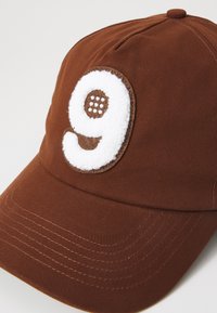Cappello marrone in cotone di forma arrotondata. Presenta una toppa bianca e pelosa con il numero "9" e accenti marroni. Dettagli cuciti sul visiera.