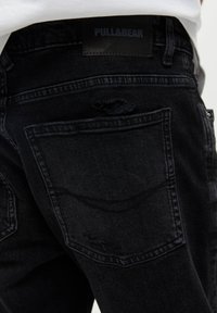 Czarne dżinsy typu denim z przetartą tylną kieszenią. Posiadają metkę Pull&Bear oraz lekko wyblakłą teksturę, co nadaje im nowoczesny, dopasowany krój.