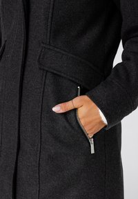 Donkergrijze wollen jas met een ritszak, uitgerust met een puntige kraag en een gladde textuur. Close-up van een hand die in de zak steekt.