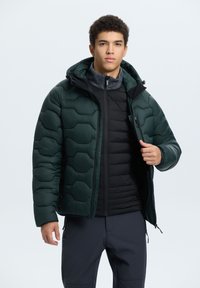 Puffer bunda, tmavě zelená, prošívaný design s kapucí. Natažená přes černou zipovou vestu. Hladký povrch, nastavitelné manžety a kapsy.