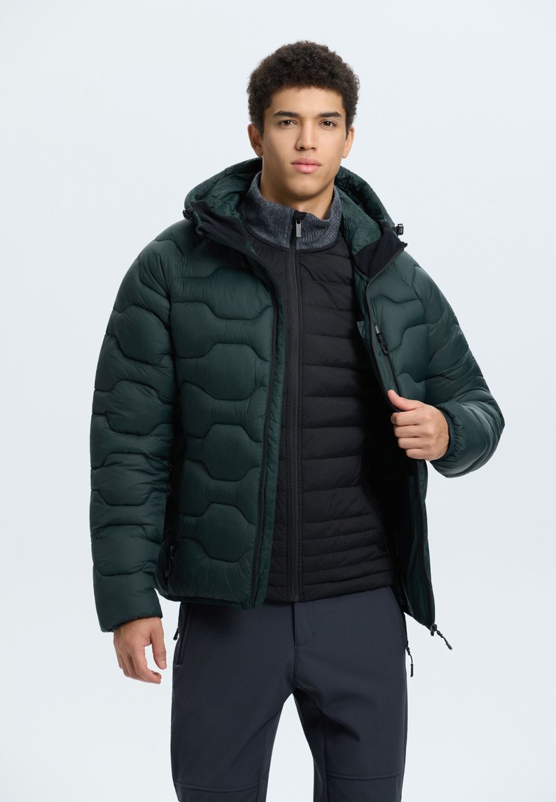 Puffer bunda, tmavě zelená, prošívaný design s kapucí. Natažená přes černou zipovou vestu. Hladký povrch, nastavitelné manžety a kapsy.