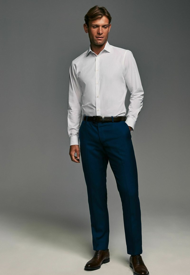 Cămașă albă cu guler deschis, pantaloni tunși de culoare bleumarin, pantofi eleganți maro și o curea neagră, stând în fața unui fundal gri.