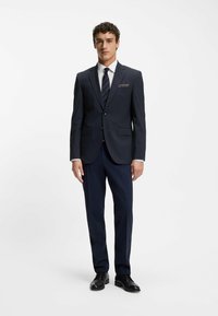 Completo blu navy con un design aderente, realizzato in tessuto liscio. Presenta un blazer monopetto e pantaloni su misura. Scarpe eleganti nere.