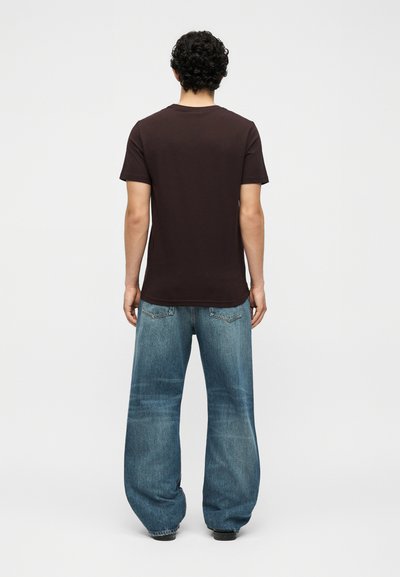 Άνδρας με μαύρα σγουρά μαλλιά στέκεται πλάτη φορώντας μια απλή σκουρόκαφέ μπλούζα και φαρδιά, ξεθωριασμένα μπλε τζιν με μαύρα παπούτσια.