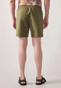 Green swim shorts with an elastic waistband, smooth texture, and side pockets. Worn with black sandals, showcasing a casual design.