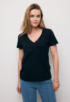Vrouw met blond haar die een zwart T-shirt met V-hals en korte mouwen draagt, gecombineerd met een blauwe spijkerbroek, tegen een effen witte achtergrond.