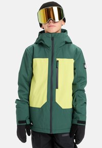 Zielono-żółta kurtka narciarska z kieszenią na zamek, elastycznymi mankietami i kapturem, w zestawie z czarnymi rękawiczkami i lustrzanymi goglami.