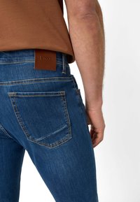 Tmavě modré džíny z denimu s hnědým koženým štítkem, na kterém je uvedeno jméno značky "Brax". Viditelná zadní kapsa a detaily šití.