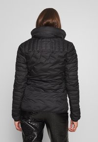 Giacca a vento nera con colletto alto, texture trapuntata e maniche lunghe, abbinata a pantaloni neri lucidi e a fantasia.