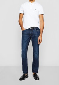 Chemise polo blanche, jeans bleu foncé, ceinture noire et chaussures noires. Coupe standard, manches courtes, texture légère sur la chemise, design classique.