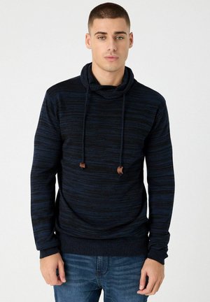 Sudadera con capucha azul marino con rayas texturizadas, cordones con detalles marrones y puños acanalados. Llevada sobre jeans de mezclilla.