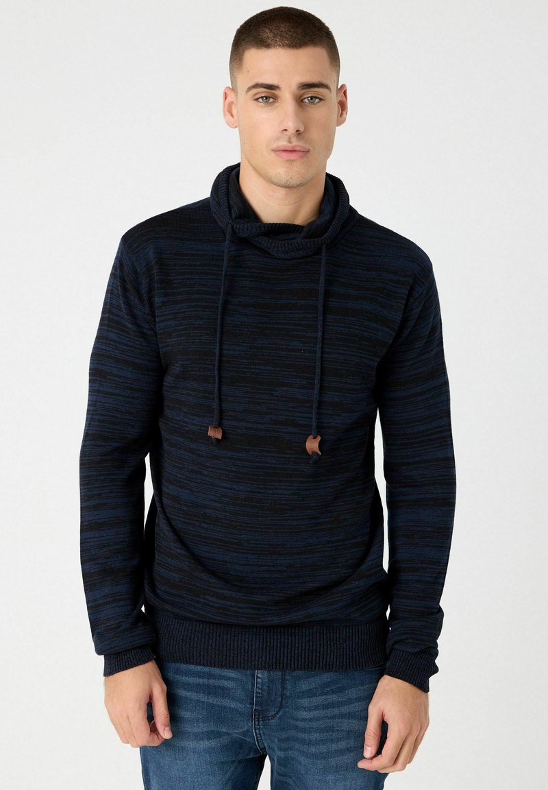 Sudadera con capucha azul marino con rayas texturizadas, cordones con detalles marrones y puños acanalados. Llevada sobre jeans de mezclilla.