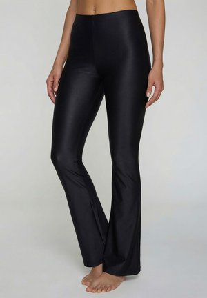 Frau trägt hochtaillierte, schwarze glänzende Schlaghosen und steht barfuß vor einem schlichten, hellen Hintergrund.