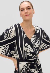 Robe noire et crème avec des motifs floraux, décolleté en V, manches amples et taille nouée, fabriquée en tissu léger, idéale pour un port décontracté.