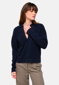 Dunkelblaue, gestrickte Pullover mit V-Ausschnitt und Kragen, ausgestattet mit langen Ärmeln und einem legeren Schnitt. Kombiniert mit beigen, karierten Hosen.