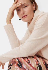 Jasnoróżowy sweter z golfem z prążkowanymi mankietami, noszony z wzorzystą spódnicą z abstrakcyjnymi kształtami w ciepłych odcieniach. Prosta biżuteria jako akcent.