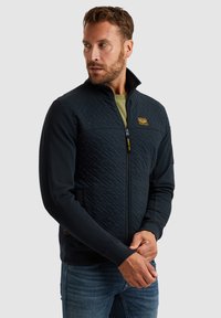 Navy-blauer, gesteppter Reißverschlussjacke mit erhöhtem Kragen und Seitentaschen, ausgestattet mit einem gelben Logopatch und strukturiertem Stoff an der Vorderseite.