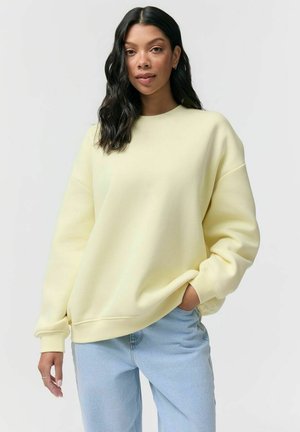 Kobieta o falujących czarnych włosach, ubrana w jasnżółtą, oversize'ową bluzę i jasnoniebieskie dżinsy, stojąca z jedną ręką w kieszeni.